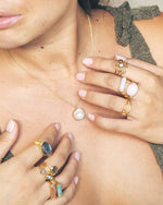 Close-up of a person wearing multiple gemstone rings on both hands, including turquoise and opal, along with a necklace featuring a pendant with a round white stone. The person is dressed in a green textured top.