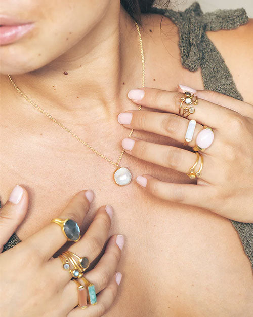 Close-up of a person wearing multiple gemstone rings on both hands, including turquoise and opal, along with a necklace featuring a pendant with a round white stone. The person is dressed in a green textured top.