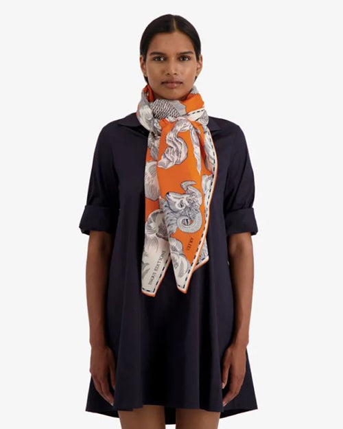 A woman wears a dark-colored dress with short sleeves that reach the elbows. The woman wears a bold orange scarf with white and grey patterns, including what appears to be stylized animal figures and decorative motifs. 