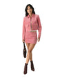 Person wearing a pink cropped jacket with long sleeves, flap pockets, and gold buttons, paired with a matching mini skirt featuring front pockets and gold buttons. The outfit is accessorized with a dark brown handbag and brown ankle boots.