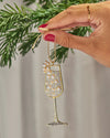 A hand holding a sparkling champagne glass ornament decorated with rhinestones and pearl accents, hanging from a gold string, with green pine branches in the background.