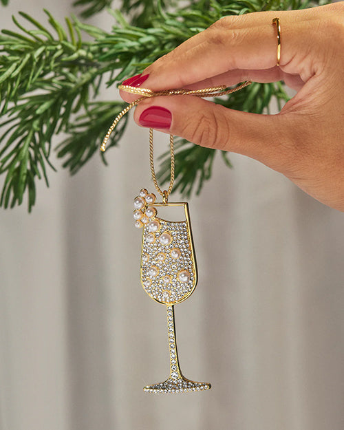 A hand holding a sparkling champagne glass ornament decorated with rhinestones and pearl accents, hanging from a gold string, with green pine branches in the background.