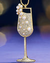 Close-up of a gold champagne glass ornament adorned with sparkling rhinestones and pearl accents, displayed against a blurred purple background.