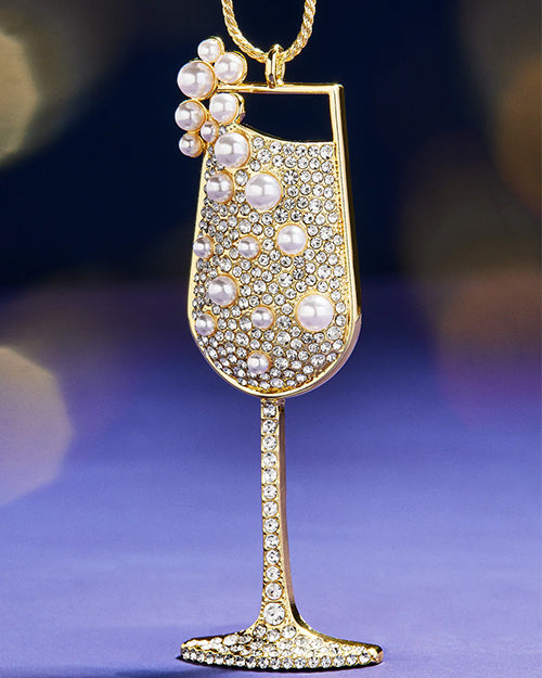 Close-up of a gold champagne glass ornament adorned with sparkling rhinestones and pearl accents, displayed against a blurred purple background.