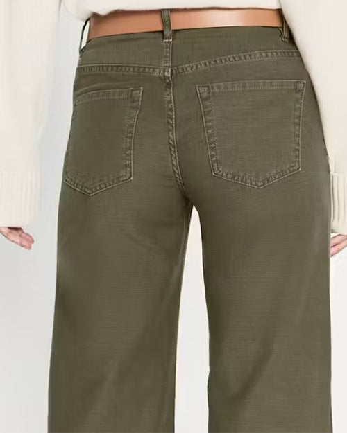 A back view of a model wearing olive green denim trousers with a wide leg fit and stitched hem. The trousers are paired with a light brown belt and white top.