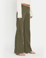 A side view of olive green denim trousers with cinching detail mid leg that starts a wide leg fit. The trousers are paired with a light brown belt, a white top, and nude open-toe sandals.