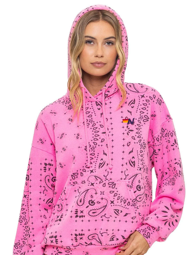 A person wearing a bright pink hoodie with a black bandana-style pattern is shown from the waist up. The hoodie features a front pocket, drawstrings, and a small multicolor logo on the left chest. The hood is up, and the person has long, wavy blond hair falling over the shoulders.