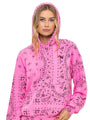 A person wearing a bright pink hoodie with a black bandana-style pattern is shown from the waist up. The hoodie features a front pocket, drawstrings, and a small multicolor logo on the left chest. The hood is up, and the person has long, wavy blond hair falling over the shoulders.