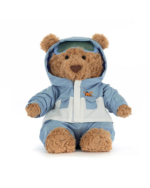 Plush teddy bear wearing a light blue and white winter outfit with a hooded jacket, embroidered orange and black helicopter logo on the chest, matching pants, and green goggles on its head.