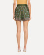 A backside, waist down view of a model wearing a bright yellow top tucked into high-waisted green shorts that feature a vibrant floral pattern with vertical stripe designs and horizontal bands around the leg openings and a wide belt made of the same fabric, tied in a large bow at the waist. The person is also wearing silver open-toe high-heeled shoes, standing against a white background.