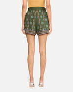 A backside view of a person wearing a bright yellow top tucked into high-waisted green shorts that feature a vibrant floral pattern. The pattern features vertical stripe designs and horizontal bands around the leg openings. The shorts have a wide belt made of the same fabric, tied in a large bow at the waist. The person is also wearing silver open-toe high-heeled shoes.