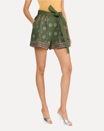 A person is wearing a bright yellow top tucked into high-waisted green shorts that feature a vibrant floral pattern. The pattern features vertical stripe designs and horizontal bands around the leg openings. The shorts have a wide belt made of the same fabric, tied in a large bow at the waist. The person has their hand in a side pocket. The person is also wearing silver open-toe high-heeled shoes.