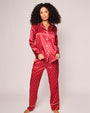 A person wearing a red  silk pajama set with white art noveaux print, a pattern of small white palm-like motifs, and white piping. The pajama set includes a button-up shirt and matching pants. 