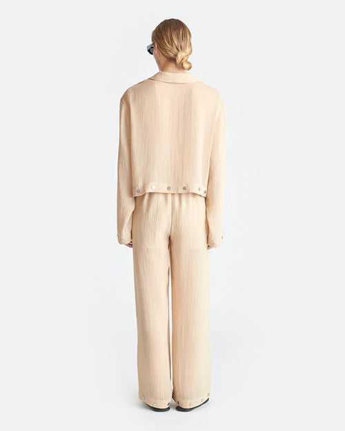 The backside view of a person wearing a beige, long-sleeved, shirt with a collar and matching flowy pants. The shirt has buttons across the waist, and the pants have buttons around the outside of the leg holes. The person's is wearing black sunglasses and sandals to match.