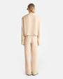 The backside view of a person wearing a beige, long-sleeved, shirt with a collar and matching flowy pants. The shirt has buttons across the waist, and the pants have buttons around the outside of the leg holes. The person's is wearing black sunglasses and sandals to match.