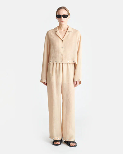 A person wearing a beige, long-sleeved, button-up shirt with a collar and matching flowy pants. The shirt has buttons down the front and at the cuffs and across the waist. The person's is wearing black sunglasses to match.