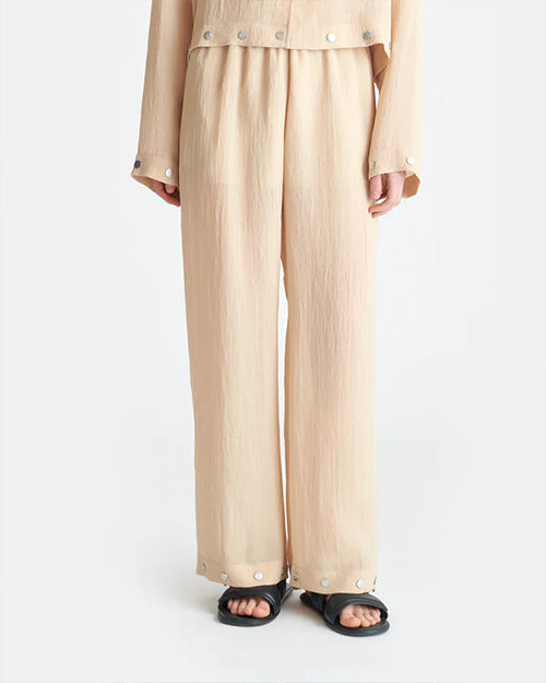 A person wearing a beige, long-sleeved shirt and matching flowy pants. The shirt has buttons at the cuffs and across the waist, and the pants have buttons around the outside of the leg holes. The person's is wearing black sandals to match.