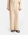 A person wearing a beige, long-sleeved shirt and matching flowy pants. The shirt has buttons at the cuffs and across the waist, and the pants have buttons around the outside of the leg holes. The person's is wearing black sandals to match.