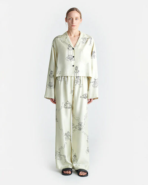 A person is seen wearing a matching pajama set consisting of a long-sleeve button-up shirt and pants. The pajama set is off-white with black line drawings of various whimsical characters and scenes such as warriors on horses and towers. The background is plain white.