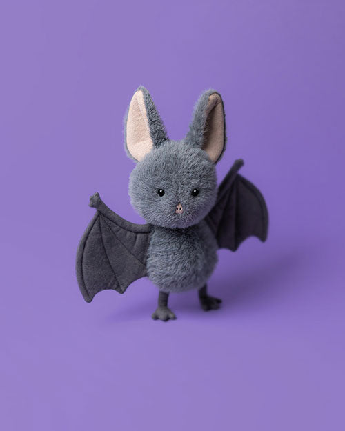 Plush toy bat with a fluffy grey body, large light pink inner ears, small black eyes, and dark grey structured wings, set against a solid purple background.
