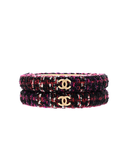 A stack of two Chanel bracelets. The bracelets feature a tweed pattern with dominant shades of pink, purple, and black. In the center of each bracelet is the iconic Chanel double ‘C’ logo in gold-tone metal, creating a striking contrast against the textured background of the tweed fabric. The bracelets are designed with a flat profile and appear to be rigid, suggesting they are bangles.