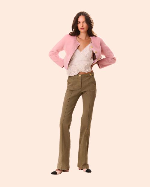 A person standing against a light-colored background, wearing olive green trousers, a white lace top, and a pink blazer. The person has one hand on their hip and is wearing black shoes with pointed toes.