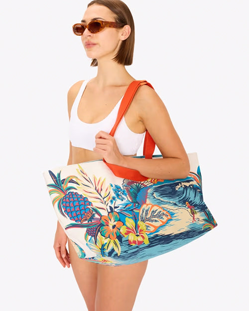 An angled view of a brunette model in profile view holding a large, colorful beach bag with tropical designs including a landscape illustration of a beach and volcano. The person is wearing a white top and brown sunglasses, standing against a white background.