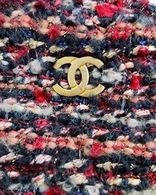  The image depicts a close-up texture of multicolored tweed fabric with red, blue, black, white and additional color threads. In the center, there’s a gold-colored metal logo resembling two interlocking letter Cs, which is associated with the luxury fashion brand Chanel. The image showcases intricate textile patterns and brand identification. 