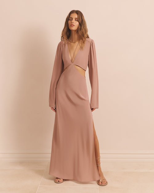 A model wearing a long-sleeved, floor-length pale blush dress with cut-out details at the waist and low V-neck. The dress has a flowing fit, and the person is wearing brown sandals.