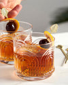 Two textured glasses filled with amber cocktails, garnished with orange peel and cherries on picks. A hand adjusts one pick, evoking a refined ambiance.