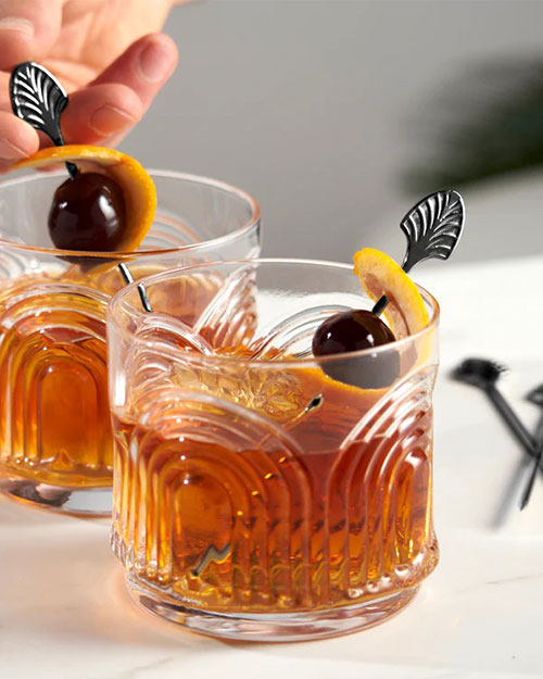 Two textured glasses filled with amber cocktails, garnished with orange peel and cherries on picks. A hand adjusts one pick, evoking a refined ambiance.