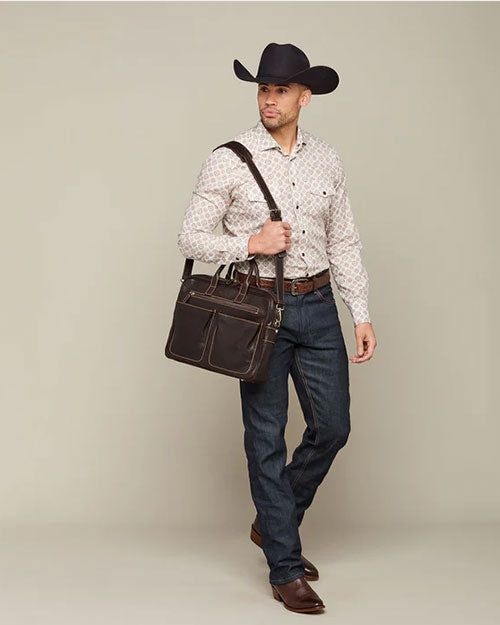 A person stands against a neutral background, wearing a cowboy hat, patterned shirt, jeans, and cowboy boots. The person is holding a dark brown leather bag and has a strap over one shoulder.