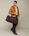 A person carrying a large brown leather duffel bag over their right shoulder. The individual wears a brown suede jacket, a white patterned shirt, dark blue jeans, and brown leather shoes. The background is a neutral solid color, providing contrast with the subject.