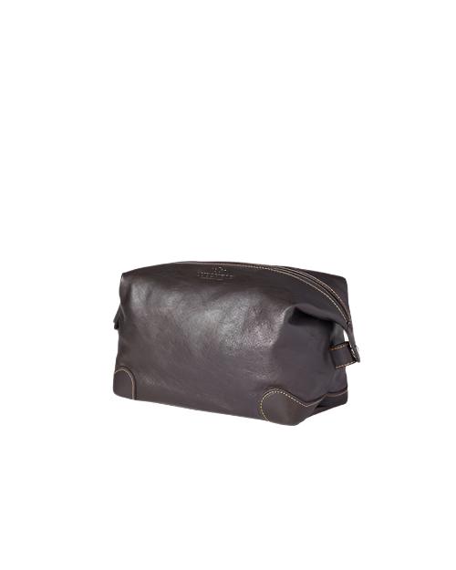 Side view of a brown leather dopp kit with a zipper closure and side strap loops, placed against a light grey background. The kit appears cylindrical with a flat base, featuring visible stitching and reinforced corners.