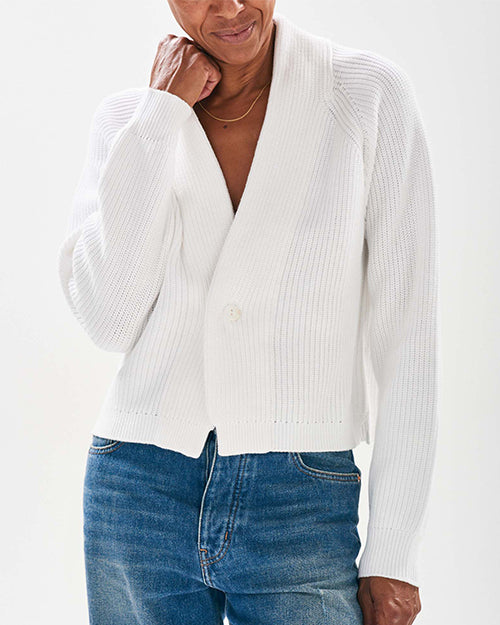 A person is wearing a white, ribbed cardigan with long sleeves, ribbed detailing a single button closure at the front. The cardigan is paired with blue jeans. 