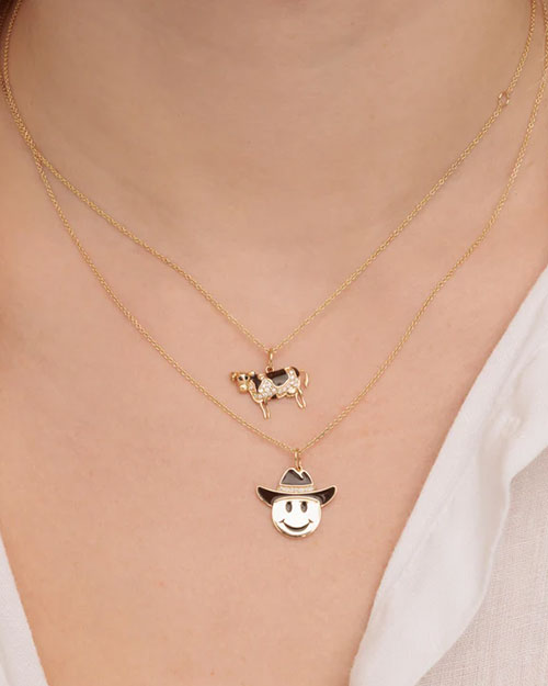 Two layered gold necklaces. The upper necklace has a cow-shaped pendant with black and white enamel, and the lower necklace features a smiley face wearing a cowboy hat with enamel detailing.