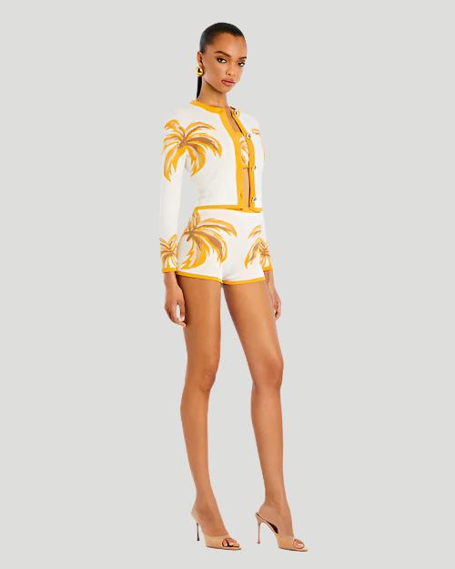 A person wearing a white and yellow cropped cardigan with a palm leaf pattern and matching high-waisted shorts and crop top, and  nude heels to complete the look.