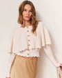 Person wearing a white ribbed knit sweater with long sleeves, scalloped hem, gold front buttons, and a cape-like shoulder layer, paired with a beige skirt. 