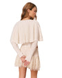 Back view of a person wearing a cream-colored knitted top with long sleeves and a cape-like overlay, paired with a lace skirt. The hands are clasped behind the back, and the face is turned to the side.