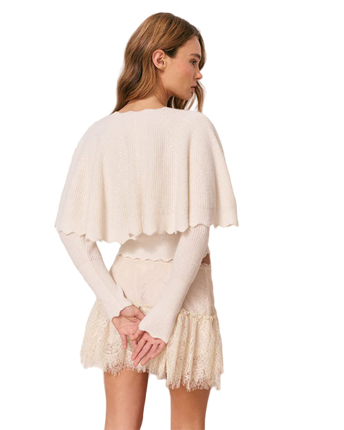 Back view of a person wearing a cream-colored knitted top with long sleeves and a cape-like overlay, paired with a lace skirt. The hands are clasped behind the back, and the face is turned to the side.