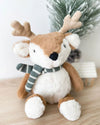 A plush reindeer doll wearing a striped scarf sits on a wooden surface. Behind it, a small snowy tree and house evoke a cozy, festive atmosphere.