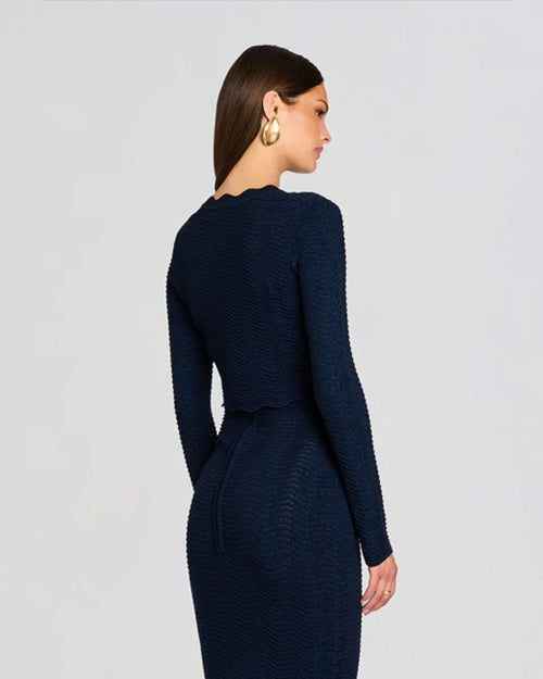A knee-up backside view of a brunette model with her hair tucked to the side wearing a large round gold earring, and a dark blue matching outfit consisting of a long-sleeved cardigan with a ribbed design and a fitted dress. The background is plain white.