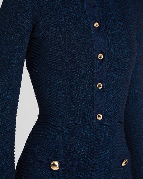 A close-up torso shot of a model wearing a dark blue matching outfit consisting of a long-sleeved cardigan with a ribbed design and gold buttons down the front, and a skirt including two pockets with gold buttons by the waist. 