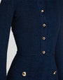 A close-up torso shot of a model wearing a dark blue matching outfit consisting of a long-sleeved cardigan with a ribbed design and gold buttons down the front, and a skirt including two pockets with gold buttons by the waist. 