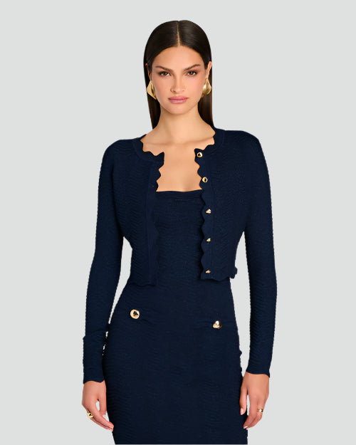 A knee-up shot of a brunette model wearing large round gold earrings, and a dark blue matching outfit consisting of a long-sleeved cardigan with scalloped edges and gold buttons, and a dress with two gold buttons by the waist. The background is plain white.
