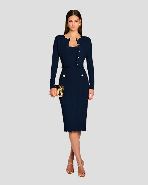 A full body shot of a brunette model wearing large round gold earrings, and a dark blue matching outfit consisting of a long-sleeved cardigan with scalloped edges and gold buttons, and a dress with two gold buttons by the waist. She is also holding a gold clutch and wearing open-toed gold heels. The background is plain white.
