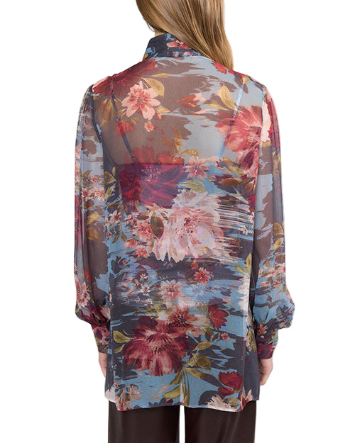 Back view of a person wearing a sheer, long-sleeved blouse with a floral pattern in red, pink, white, and yellow, set against a dark, translucent background with hints of blue. The blouse has a relaxed fit with slightly puffed sleeves gathered at the cuffs. The person has long, light brown hair and is also wearing dark-colored pants.