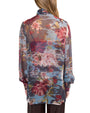 Back view of a person wearing a sheer, long-sleeved blouse with a floral pattern in red, pink, white, and yellow, set against a dark, translucent background with hints of blue. The blouse has a relaxed fit with slightly puffed sleeves gathered at the cuffs. The person has long, light brown hair and is also wearing dark-colored pants.