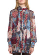 A person wearing a long-sleeved blouse made of sheer fabric with a vibrant floral pattern in red, blue, pink, and white. The blouse features vertical pleats down the front, a short stand-up collar, and slightly puffed cuffs. Buttons run through the center, adding structure to the flowing, semi-transparent design.