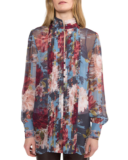 A person wearing a long-sleeved blouse made of sheer fabric with a vibrant floral pattern in red, blue, pink, and white. The blouse features vertical pleats down the front, a short stand-up collar, and slightly puffed cuffs. Buttons run through the center, adding structure to the flowing, semi-transparent design.